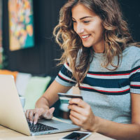 A woman holding a credit card in one hand while the other hand is using a laptop. 