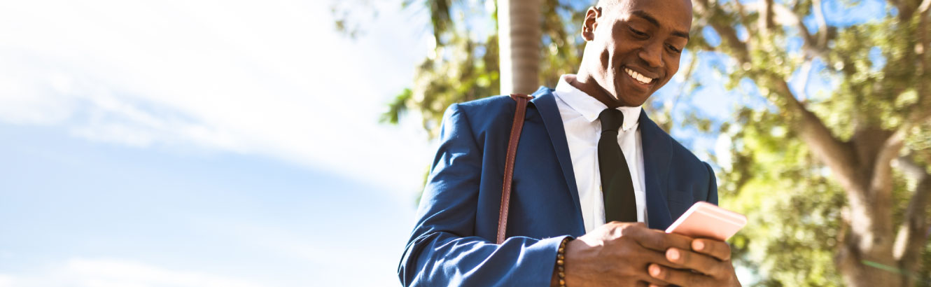 A young businessman in a suit walking outside looking down at his phone smiling. 